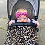 baby, blue_eyes, headband, pink_headband, stroller, blanket, leopard_print, outdoor, sidewalk, infant, child, cute, person, young_child, hand, clothing, urban, seat, safety_bar, expression