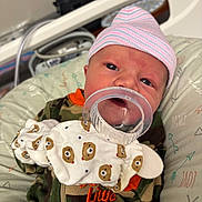 Oakley joined the competition — help win amazing prizes! baby, newborn, infant, hat, pacifier, mitten, bear_pattern, camo_clothing, cushion, patterned_bedding, face, person, cute, resting, closeup, indoors, child, pink_hat, orange_text, soft_lighting
