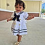 toddler, child, girl, dress, sailor_dress, white_dress, golden_shoes, bows, sidewalk, outdoor, walking, short_hair, curly_hair, smiling, cute, person, young_child, sunlight, building, concrete