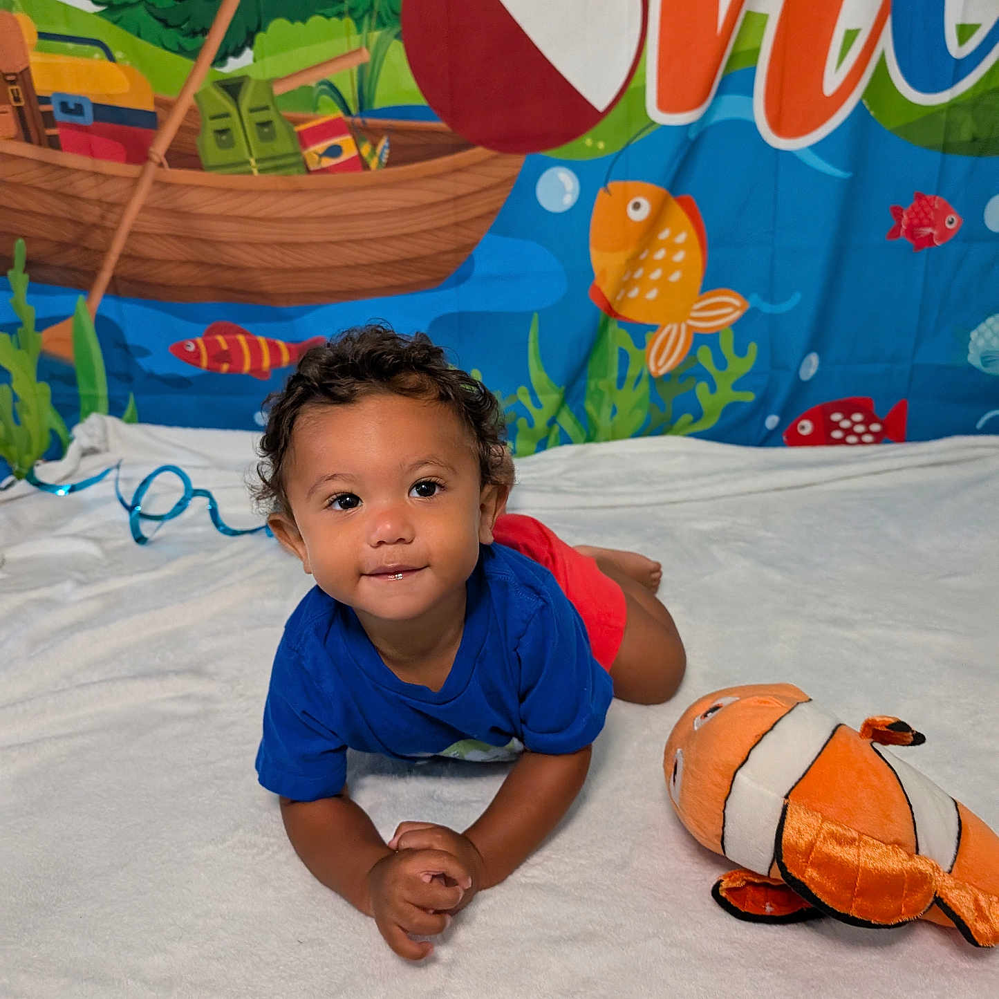 Marcel joined the competition — help win amazing prizes! blue_shirt, boat, child, clownfish, colorful_banner, curly_hair, cute, fish, floor_rug, happy, indoor, playing, plush_toy, portrait, red_shorts, smiling, toddler, underwater_background, water_splash, white_blanket