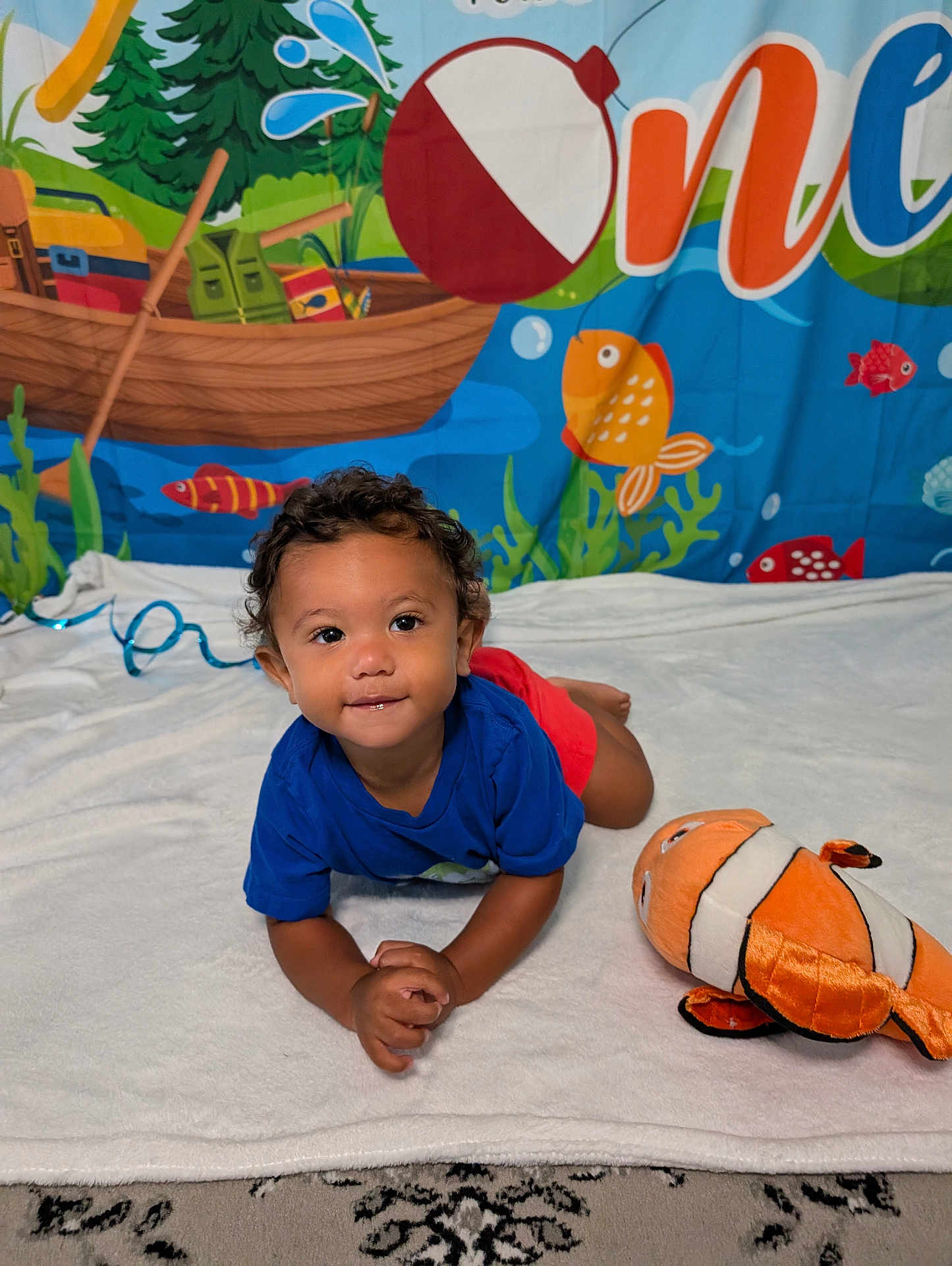 Marcel joined the competition — help win amazing prizes! toddler, child, blue_shirt, red_shorts, curly_hair, smiling, plush_toy, clownfish, white_blanket, underwater_background, colorful_banner, fish, boat, water_splash, indoor, playing, cute, portrait, happy, floor_rug