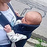 baby, child, adult, baby_carrier, outdoor, sidewalk, street, clothing, person, infant, holding, residential_area, grass, crack, road, casual_wear, head, face, young_adult, daytime