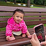 baby, child, pink_jacket, polka_dots, bench, park, outdoor, smiling, cute, curly_hair, person, photography, smartphone, hands, nature, trees, greenery, sitting, happy, portrait