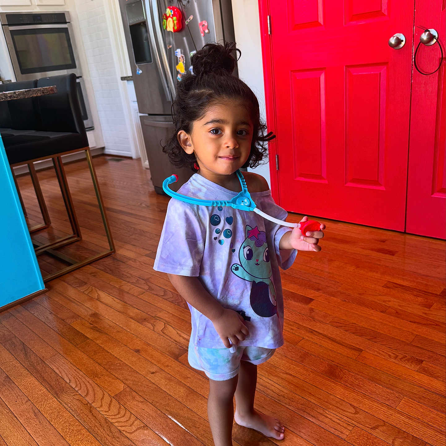 Leena is registered to the contest to win money with this photo: barefoot, cabinet, cartoon_character, child, hair_bun, home, indoor, kitchen, oven, person, playful, purple_shirt, red_door, refrigerator, smiling, standing, stethoscope, toddler, toy, wooden_floor
