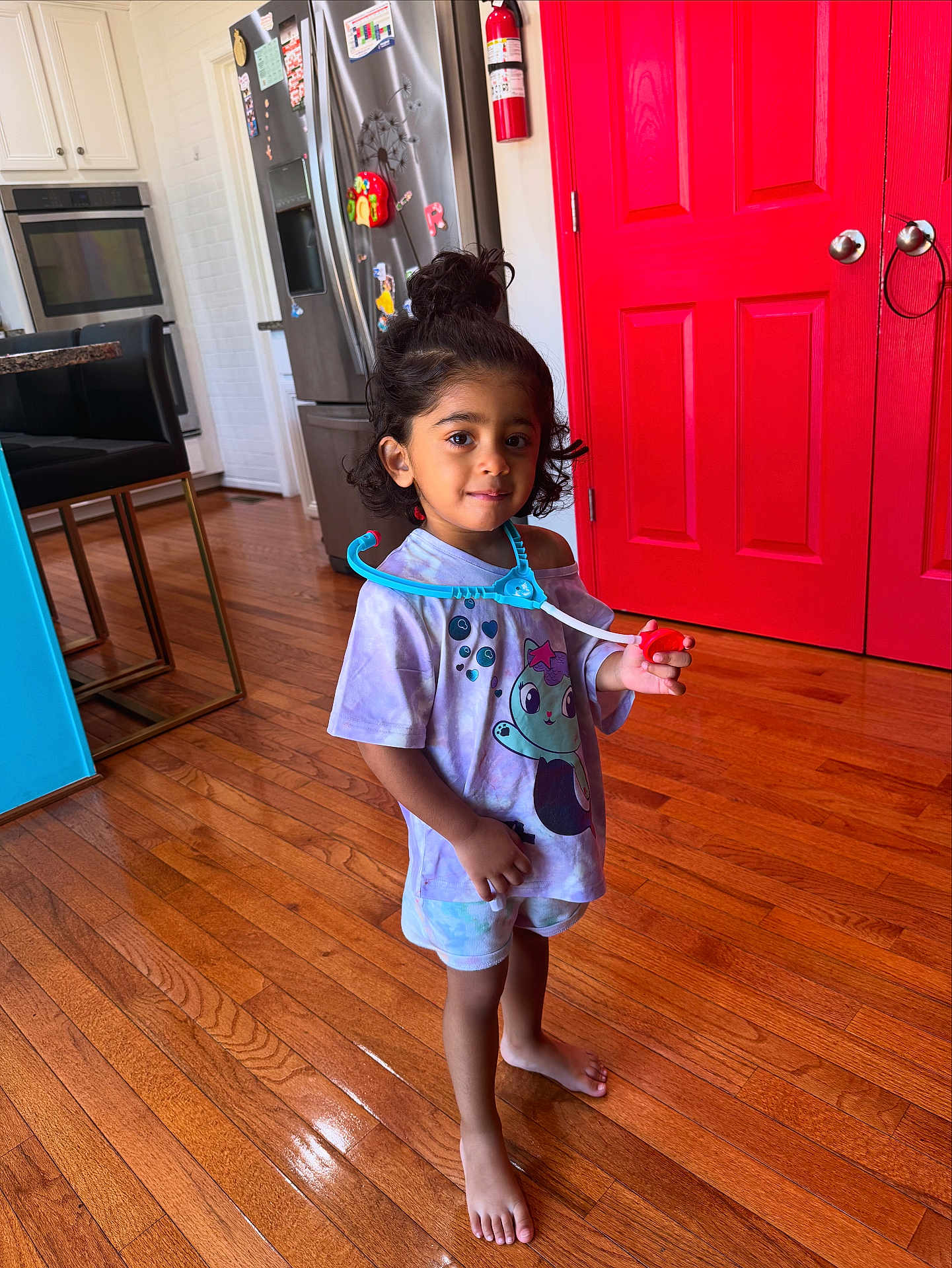 Leena is registered to the contest to win money with this photo: toddler, child, barefoot, toy, stethoscope, purple_shirt, cartoon_character, wooden_floor, red_door, kitchen, refrigerator, oven, cabinet, hair_bun, indoor, playful, person, smiling, standing, home