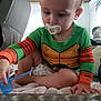 Oakley is registered to the contest to win money with this photo: toddler, child, pacifier, green_clothing, orange_sleeves, muffin_tin, blue_scoop, diaper, indoor, carpet, furniture, play, curious, young_child, baby, sitting, hand, face, light, room