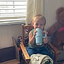 toddler, child, chair, blue_cup, blanket, pacifier, window, curtain, wooden_furniture, teddy_bear, floor, indoor, smiling, clothing, jeans, green_shirt, toy, cardboard_box, natural_light, home