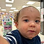 toddler, child, face, curly_hair, blue_shirt, striped_sleeve, indoor, store, shopping_cart, big_eyes, close_up, young_child, cute, expression, person, portrait, retail, curiosity, baby, casual_clothing