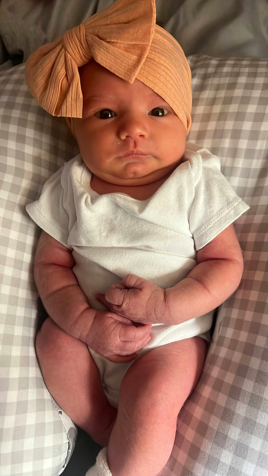 Elizabeth is registered to the contest to win money with this photo: baby, infant, headband, bow, white_onesie, pillow, checkered_pattern, hands, legs, skin, face, eyes, indoor, newborn, cute, resting, clothing, soft_light, portrait, child