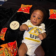 Justice is registered to the contest to win money with this photo: baby, black_blanket, cheetos, cute, fabric, indoor, infant, lying_down, necklace, onesie, orange, person, pillow, portrait, sign, snack_bags, text, white_clothing, wide_eyes, young_child