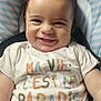 baby, smiling, child, tshirt, text, striped_cushion, infant, happy, portrait, seated, clothing, cute, indoors, face, person, sitting, young_child, joyful, casual_wear, closeup