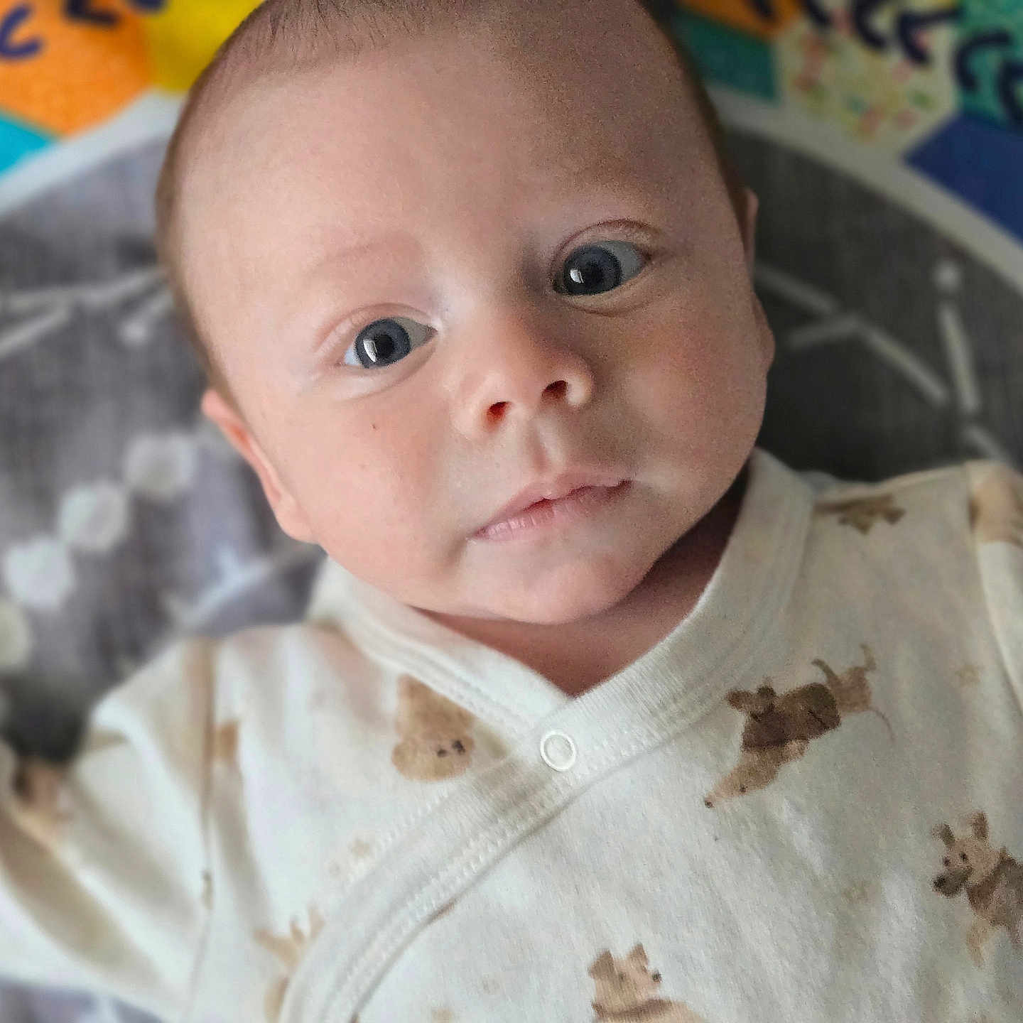 Orion is registered to the contest to win money with this photo: animal_print, baby, child, clothing, colorful, cute, expression, eyes, face, head, indoors, infant, lying_down, newborn, onesie, person, play_mat, portrait, skin, soft