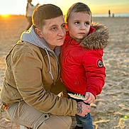 Noah participe au concours pour gagner de l'argent avec cette photo : adult, child, beach, sunset, sand, feather, jacket, hood, tattoo, crouching, holding, outdoor, warm_clothing, casual, portrait, two_people, nature, expression, footwear, family