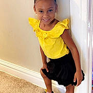 Deasia is registered to the contest to win money with this photo: child, girl, smiling, portrait, indoor, standing, yellow_shirt, black_skirt, sneakers, hair_bow, flowers, curly_hair, doorway, wall, carpet, home, fashion, playful, full_body, natural_light