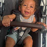 Jacob is registered to the contest to win money with this photo: toddler, child, stroller, smiling, curly_hair, safety_harness, baby, happy, seat, clothing, outdoor, person, cute, infant, legs, hand, baby_stroller, seatbelt, playful, portrait