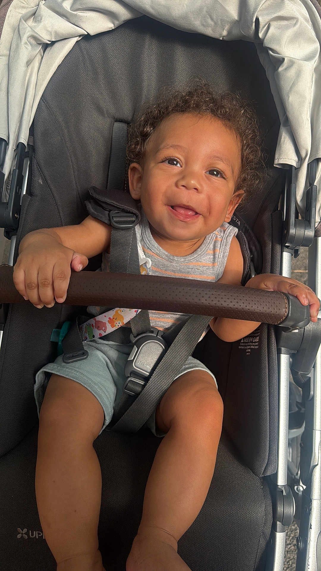 Jacob is registered to the contest to win money with this photo: toddler, child, stroller, smiling, curly_hair, safety_harness, baby, happy, seat, clothing, outdoor, person, cute, infant, legs, hand, baby_stroller, seatbelt, playful, portrait