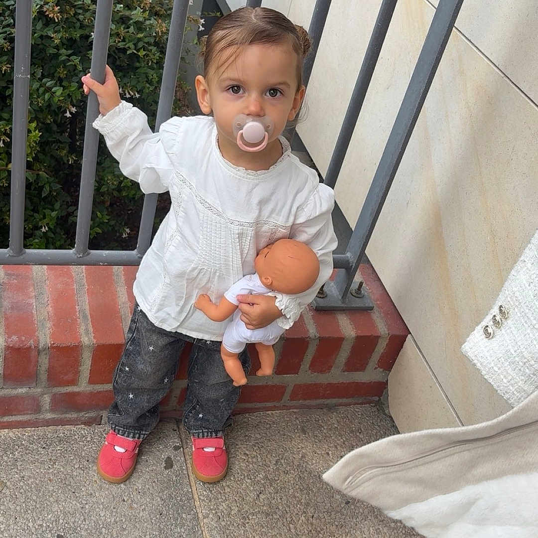Kamilla a rejoint le concours — aidez-le/la à gagner de superbes lots ! baby_doll, brick_wall, casual_clothing, child, concrete_floor, cute, doll, greenery, holding, jeans, metal_railings, outdoor, pacifier, person, portrait, red_shoes, standing, toddler, white_blouse, young_child