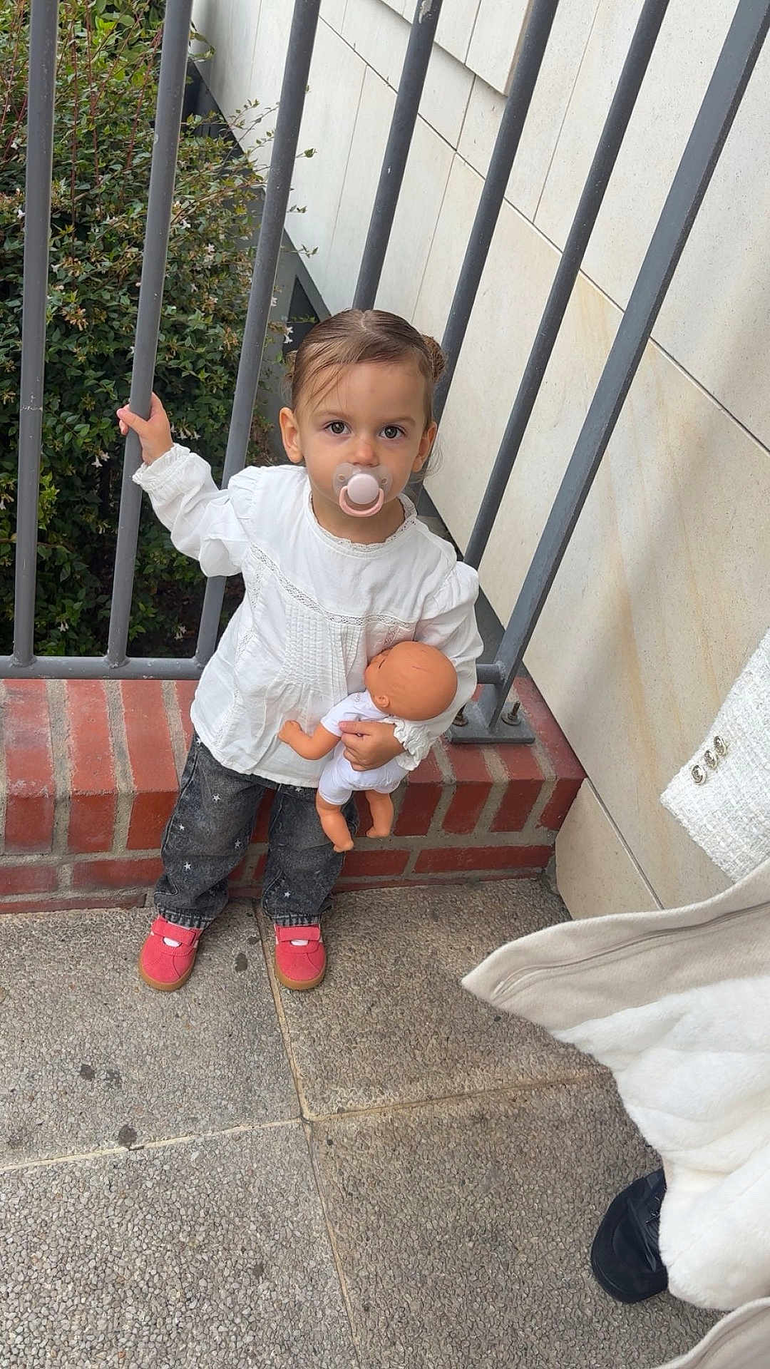 Kamilla a rejoint le concours — aidez-le/la à gagner de superbes lots ! toddler, child, pacifier, doll, white_blouse, jeans, red_shoes, metal_railings, brick_wall, concrete_floor, greenery, outdoor, person, baby_doll, holding, standing, casual_clothing, cute, young_child, portrait
