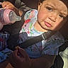 toddler, child, baby_bottle, car_seat, seatbelt, floral_clothing, sunlight, shadow, hand, face, expression, snack, young_child, indoors, seat, person, cute, portrait, closeup, child_seat