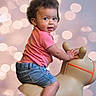 Sophia joined the competition — help win amazing prizes! toddler, child, curly_hair, pink_shirt, denim_shorts, inflatable_toy, toy_horse, indoor, bokeh_background, happy, smiling, cute, playful, person, portrait, baby, fun, sitting, looking_back, bright
