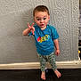 child, toddler, barefoot, blue_tshirt, patterned_pants, standing, wallpaper, dark_floor, indoor, playful, gesture, funny_face, person, young, cute, expression, home, floor, wall, casual_clothing