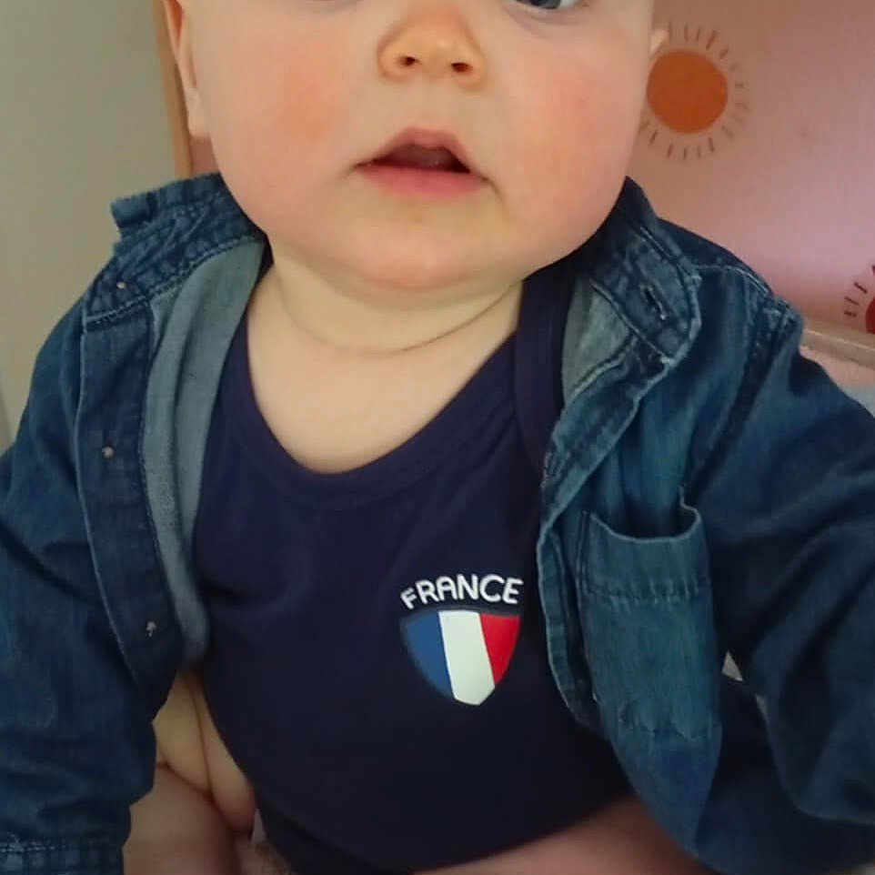 Ezio a rejoint le concours — aidez-le/la à gagner de superbes lots ! baby, blue_clothing, child, chubby_legs, curious_expression, cute, denim_jacket, face, figurines, floral_blanket, france_logo, head, indoor, infant, person, plant, portrait, room, sitting, skin