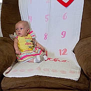 Blakely is registered to the contest to win money with this photo: armchair, baby, brown_furniture, child, colorful_clothing, cozy, curious_expression, cute, fabric, home, indoor, infant, pink_numbers, portrait, red_heart, seated, socks, soft_lighting, striped_dress, white_cloth