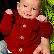 Ivan is registered to the contest to win money with this photo: baby, infant, smile, face, blue_eyes, red_outfit, cardigan, wooden_buttons, hands, adult_hand, tattoo, green_sweater, jeans, portrait, indoor, couch, close_up, cute, soft_lighting, person