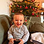 toddler, child, smiling, happy, indoor, christmas_tree, holiday, red_ornaments, couch, pillow, blanket, clothing, buttoned_shirt, blue_pants, baby_shoes, face, portrait, home, festive, cozy