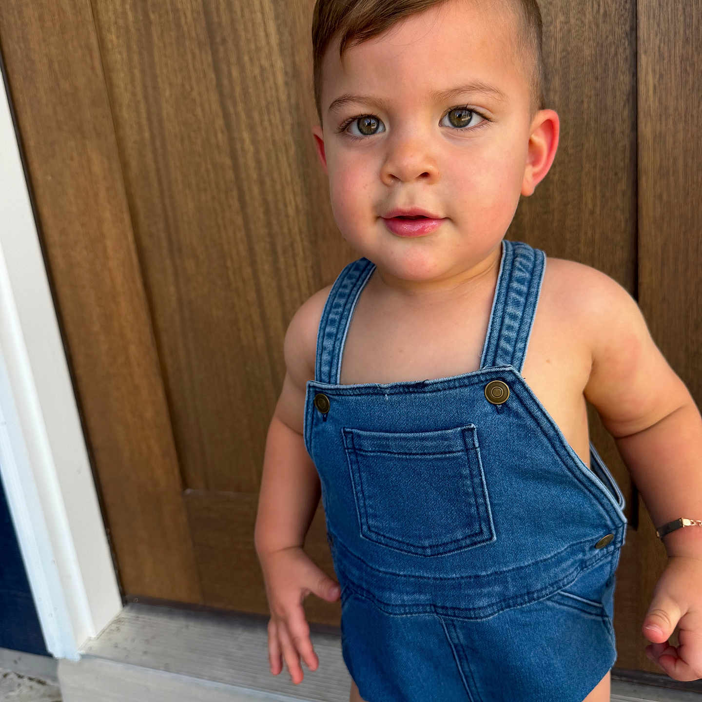 Noah Gabriel joined the competition — help win amazing prizes! toddler, child, denim_overalls, standing, door, wood, curious, face, skin, eyes, hair, outdoor, footwear_absent, expression, person, clothing, arm, hand, bracelet, young