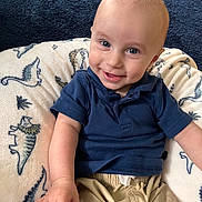 Owen is registered to the contest to win money with this photo: baby, child, smiling, blue_shirt, beige_shorts, dinosaur_pattern, cushion, indoor, portrait, happy, face, hand, sitting, infant, cute, light_skin, short_hair, clothing, background, closeup