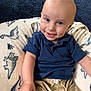 baby, child, smiling, blue_shirt, beige_shorts, dinosaur_pattern, cushion, indoor, portrait, happy, face, hand, sitting, infant, cute, light_skin, short_hair, clothing, background, closeup