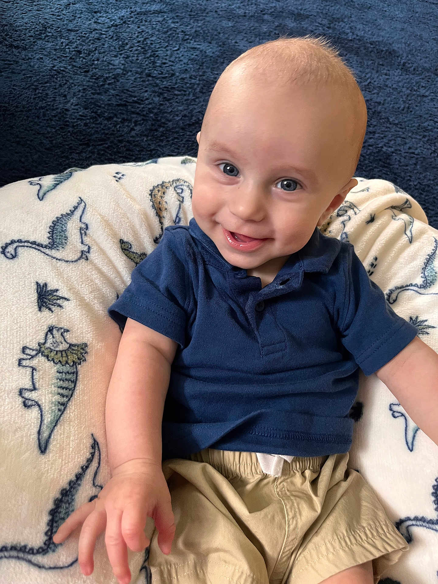 Owen is registered to the contest to win money with this photo: baby, child, smiling, blue_shirt, beige_shorts, dinosaur_pattern, cushion, indoor, portrait, happy, face, hand, sitting, infant, cute, light_skin, short_hair, clothing, background, closeup