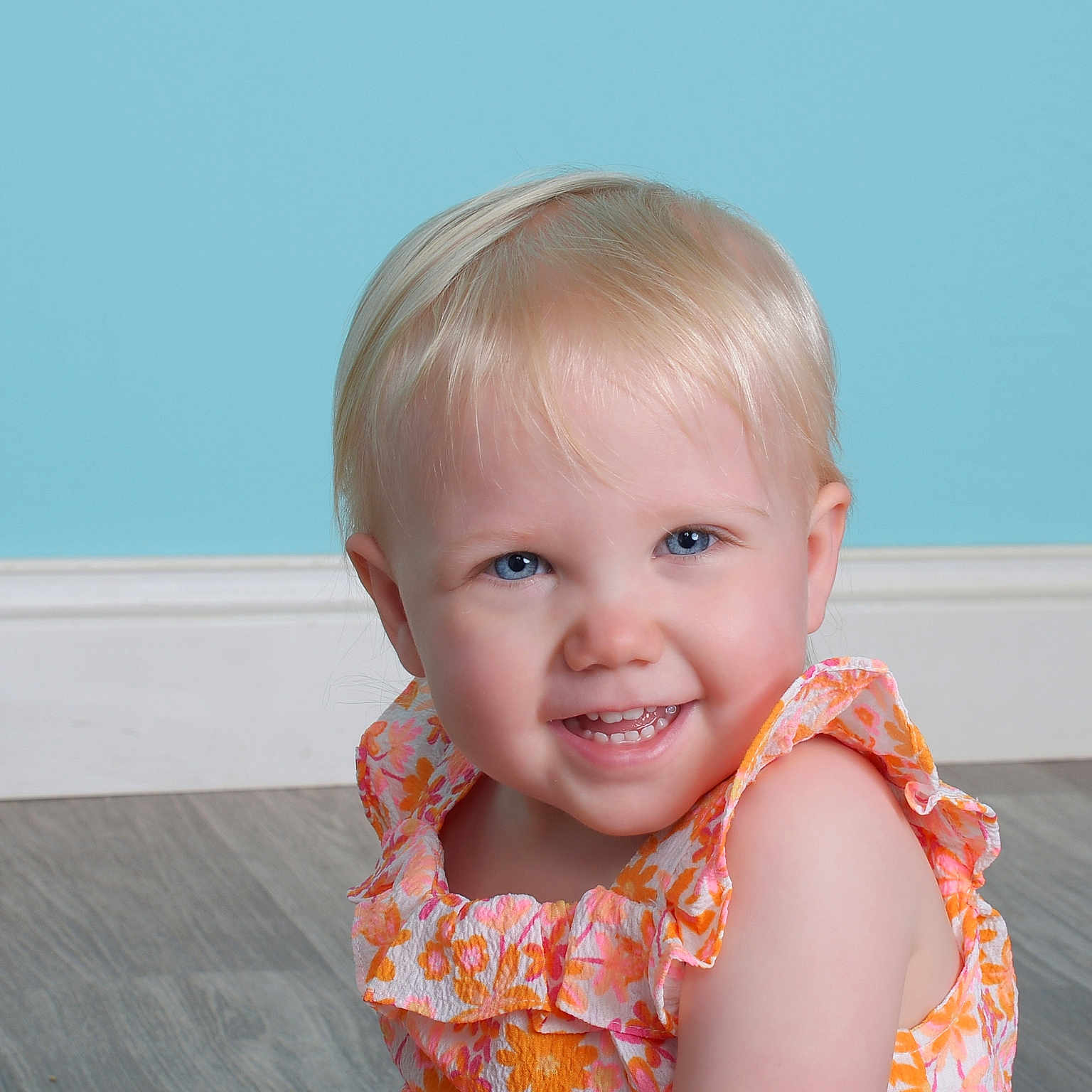 Emma is registered to the contest to win money with this photo: baby, blonde, bodypart, clothing, dress, face, finger, hair, hand, happy, hardwood, head, mouth, person, photography, plywood, portrait, sitting, smile, wood