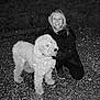 person, dog, smiling, glasses, jacket, night, gravel, outdoor, pet, curly_hair, crouching, happy, animal, friendship, canine, leash, casual, portrait, companion, black_and_white