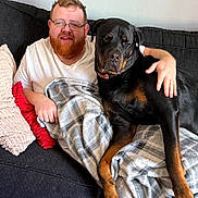 Winston joined the competition — help win amazing prizes! beard, black_dog, blanket, brown_dog, casual_clothing, comfort, couch, dog, friendship, glasses, home, indoor, man, pet, pillow, relaxing, resting, rottweiler, seated, smiling