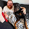 beard, black_dog, blanket, brown_dog, casual_clothing, comfort, couch, dog, friendship, glasses, home, indoor, man, pet, pillow, relaxing, resting, rottweiler, seated, smiling