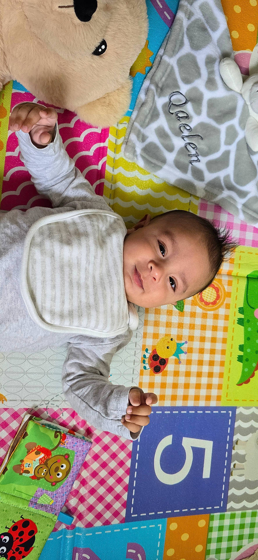 Qaelen is registered to the contest to win money with this photo: animal_print, baby, bib, blanket, child, clothing, colorful, cute, face, hand, indoors, infant, lying_down, number, pattern, person, play_mat, smile, soft_toy, toy