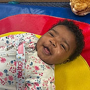 Sanai is registered to the contest to win money with this photo: baby, smiling, floral_outfit, colorful_mat, toy, plush, child, lying_down, happy, indoor, cute, person, portrait, head, face, curly_hair, playful, soft_texture, bright_colors, infant