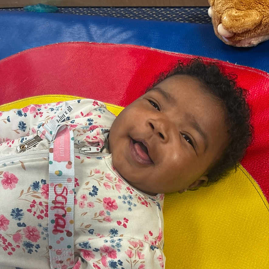 Sanai is registered to the contest to win money with this photo: baby, smiling, floral_outfit, colorful_mat, toy, plush, child, lying_down, happy, indoor, cute, person, portrait, head, face, curly_hair, playful, soft_texture, bright_colors, infant