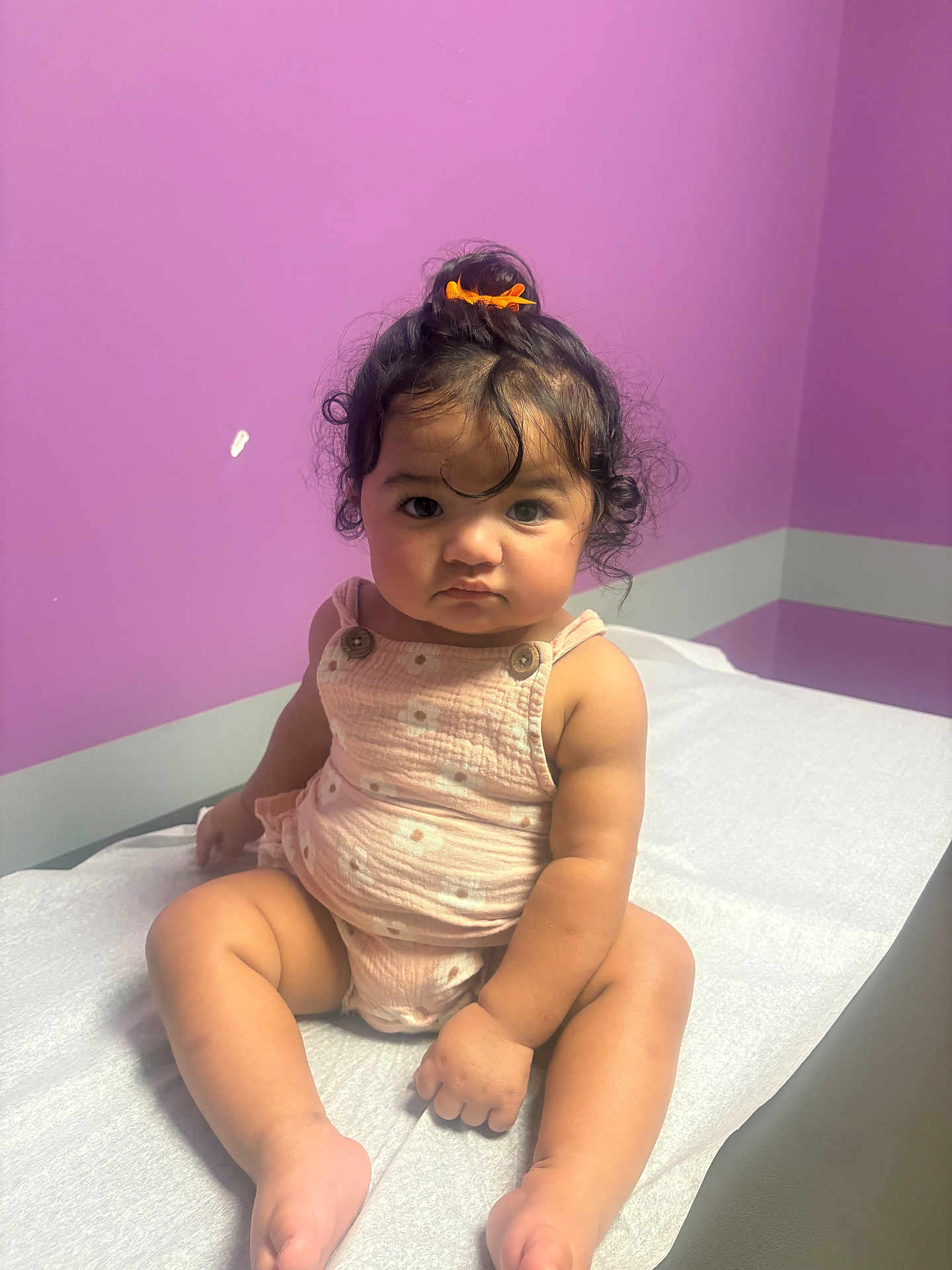 Genesis is registered to the contest to win money with this photo: baby, child, curly_hair, orange_bow, pink_romper, sitting, white_sheet, purple_wall, serious_expression, indoors, cute, chubby_cheeks, infant, portrait, young_child, skin, person, room, front_view, natural_light