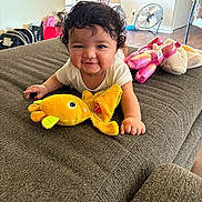 Genesis is registered to the contest to win money with this photo: baby, curly_hair, smiling, plush_toy, yellow_fish, couch, brown_fabric, indoor, living_room, fan, stuffed_animal, child, happy, person, toy, floor, table, chair, home, casual