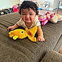 baby, curly_hair, smiling, plush_toy, yellow_fish, couch, brown_fabric, indoor, living_room, fan, stuffed_animal, child, happy, person, toy, floor, table, chair, home, casual