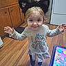 Daisy is registered to the contest to win money with this photo: child, girl, cat_ears, smiling, indoor, kitchen, wooden_floor, tablet, bottle, long_sleeve_shirt, leggings, socks, cabinet, stove, oven, towel, playful, happy, person, toy