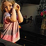 child, girl, playful, indoor, living_room, flower_vase, table, wooden_furniture, home_decor, patterned_wall, pink_shirt, casual_clothing, smiling, portrait, cute, young_child, cozy, warm_lighting, happy, person