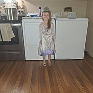 Lilly is registered to the contest to win money with this photo: child, girl, princess, crown, dress, kitchen, stove, pot, washing_machine, wooden_floor, cabinet, appliance, smile, indoors, floor, apron, long_hair, happy, standing, home