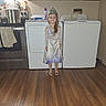 child, girl, princess, crown, dress, kitchen, stove, pot, washing_machine, wooden_floor, cabinet, appliance, smile, indoors, floor, apron, long_hair, happy, standing, home