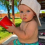 toddler, child, blue_eyes, hat, cap, red_cup, shorts, dinosaur_print, outdoor, backyard, grass, fence, patio, hand, sunny, summer, cute, baby, portrait, sitting