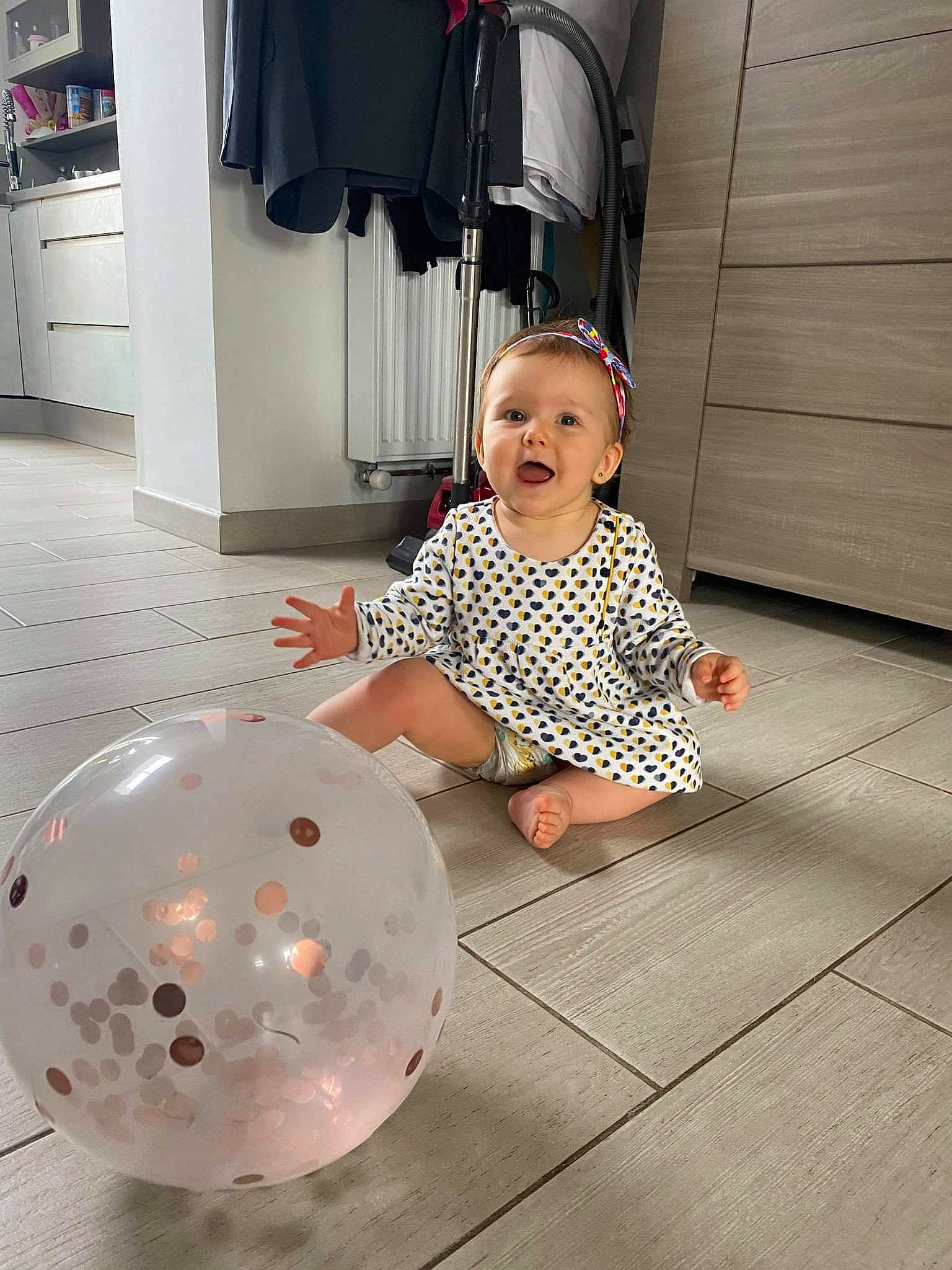 Romy participe au concours pour gagner de l'argent avec cette photo : art, baby, baby_toddler_clothing, ball, balloon, cabinetry, circle, event, floor, flooring, fun, happy, hardwood, party_supply, pattern, person, sleeve, smile, standing, toddler