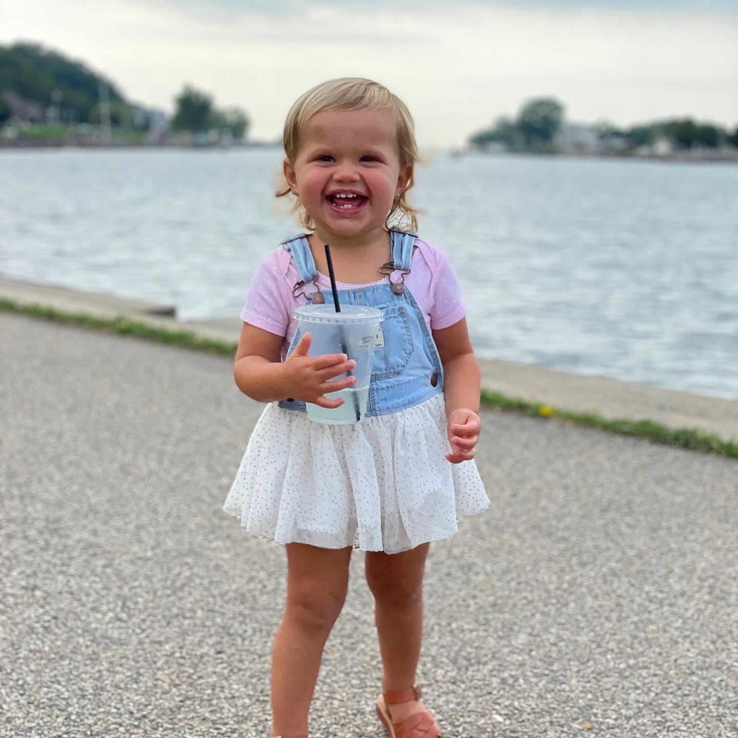 Elly May is registered to the contest to win money with this photo: blonde_hair, body_of_water, child, cup, daytime, denim, dress, happy, nature, outdoor, overalls, path, portrait, sandals, short_hair, smiling, straw, summer, toddler, water